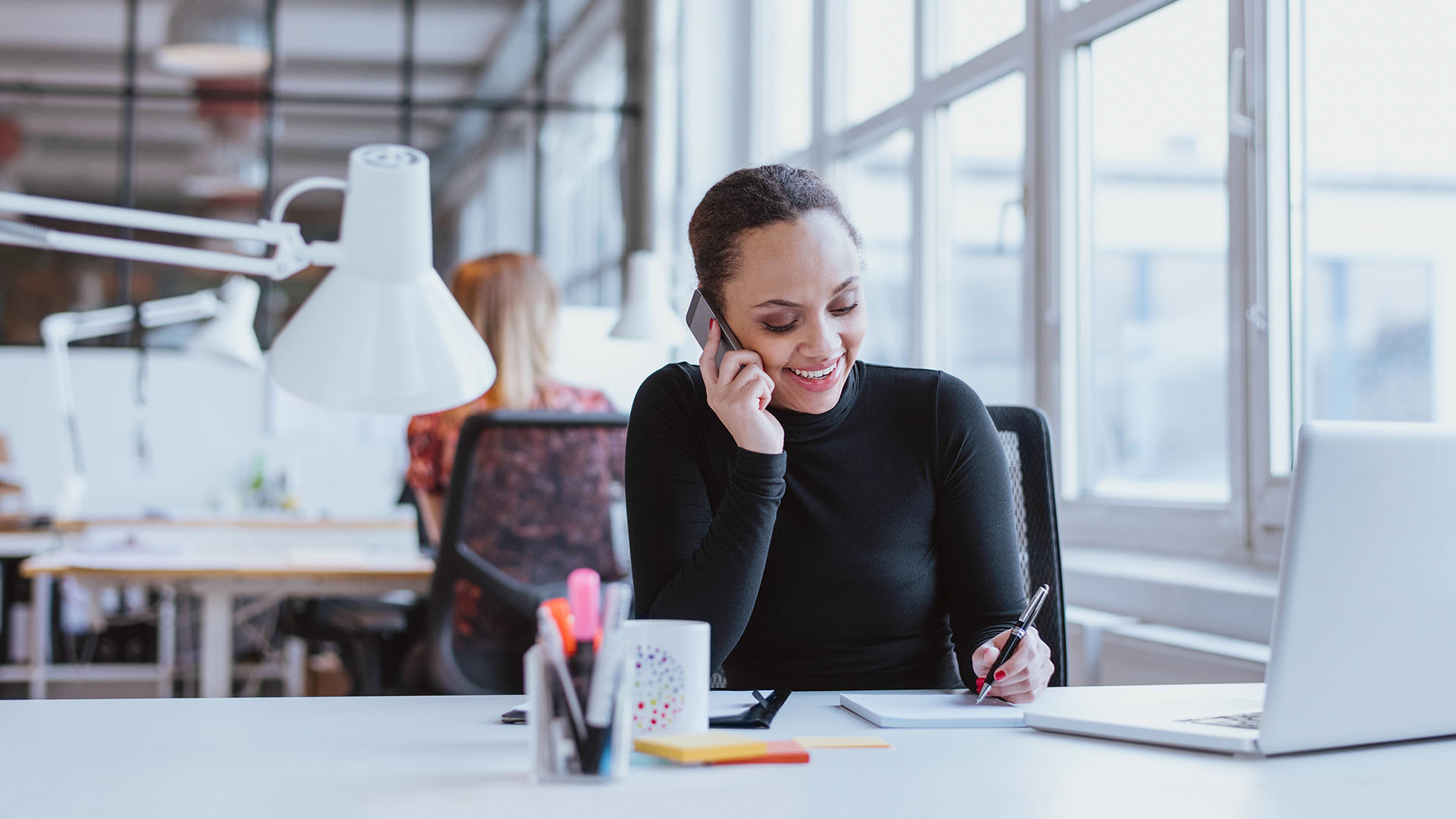 Jonge vrouw aan het bureau aan het bellen en kijken naar haar laptop